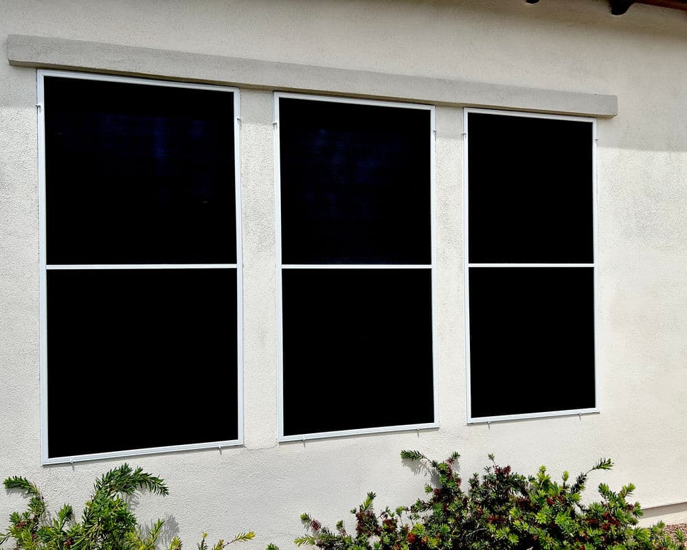 Black window shades installed on a light-colored exterior wall with green plants below.