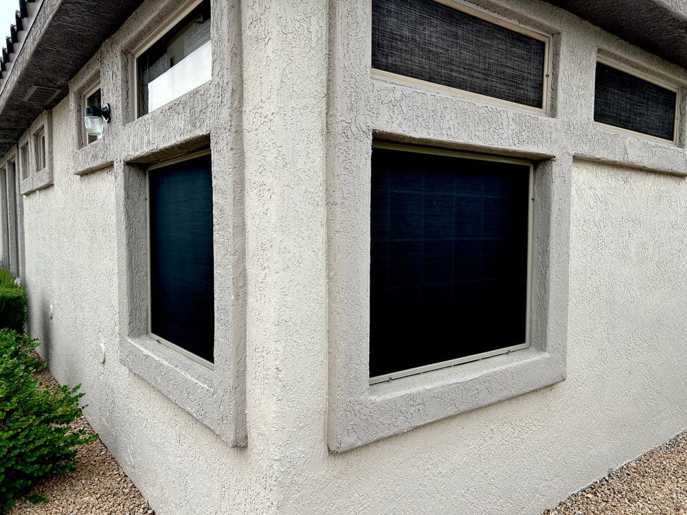 Exterior view of a textured beige wall with corner windows and dark screens, surrounded by gravel.